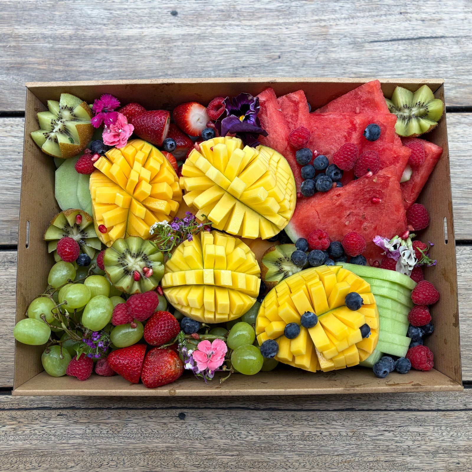 A catering box filled with assorted fresh fruit, including sliced mango, watermelon, kiwi, grapes, strawberries, blueberries, and raspberries, arranged on a weathered wooden surface.